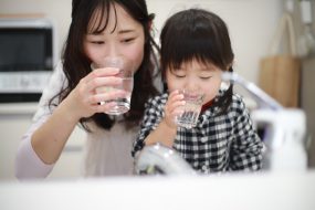 Asian mother and daughter drink glasses of water at a stainless steel sink,