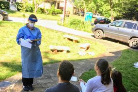 Woman dressed in personal protective equipment stands on sidewalk to ask questions to three people on their front porch.   