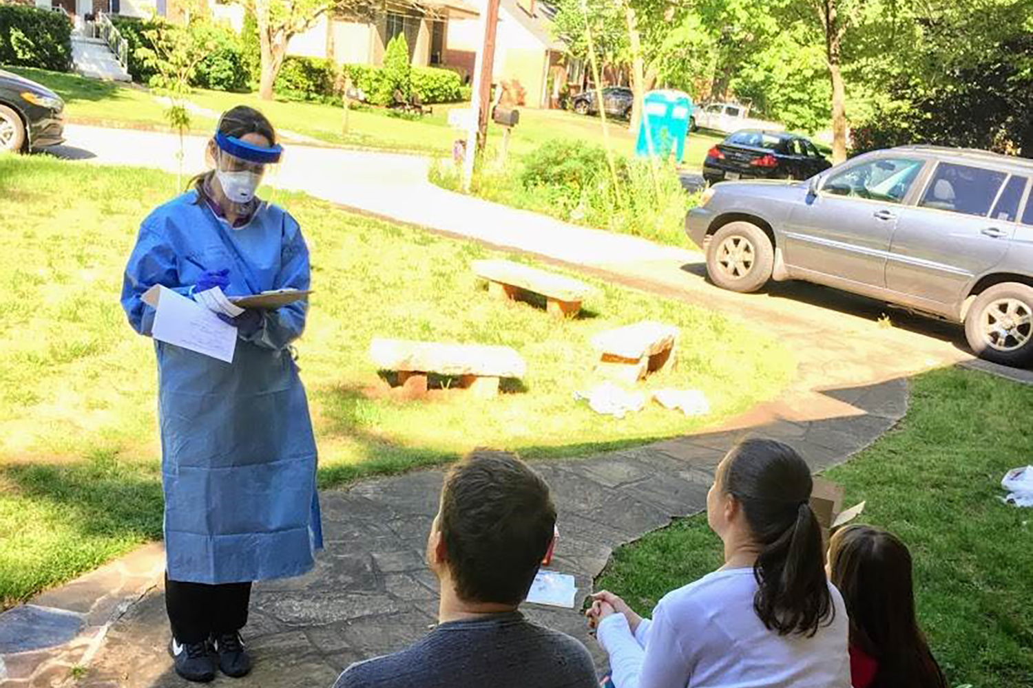 Woman dressed in personal protective equipment stands on sidewalk to ask questions to three people on their front porch.