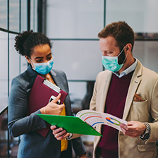 Two people with masks on and wearing business clothes examining a clip board.