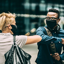 A blonde woman with short hair, a tote bag, and mask on elbow-bumping a black man who is wearing a mask and glasses.