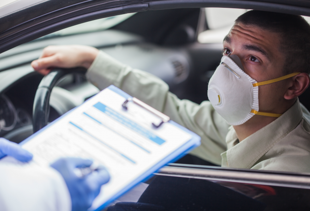 Person with N95 mask has hand on the steering wheel in a car looking up at someone is holding a clipboard.
