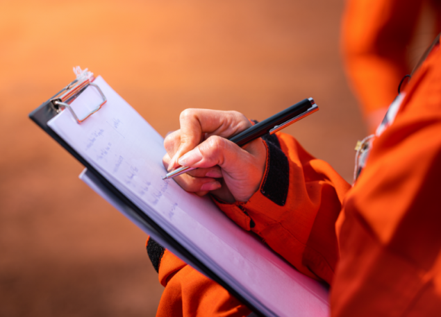 Person in orange jacket writes on a white paper on a clipboard with a black pen.