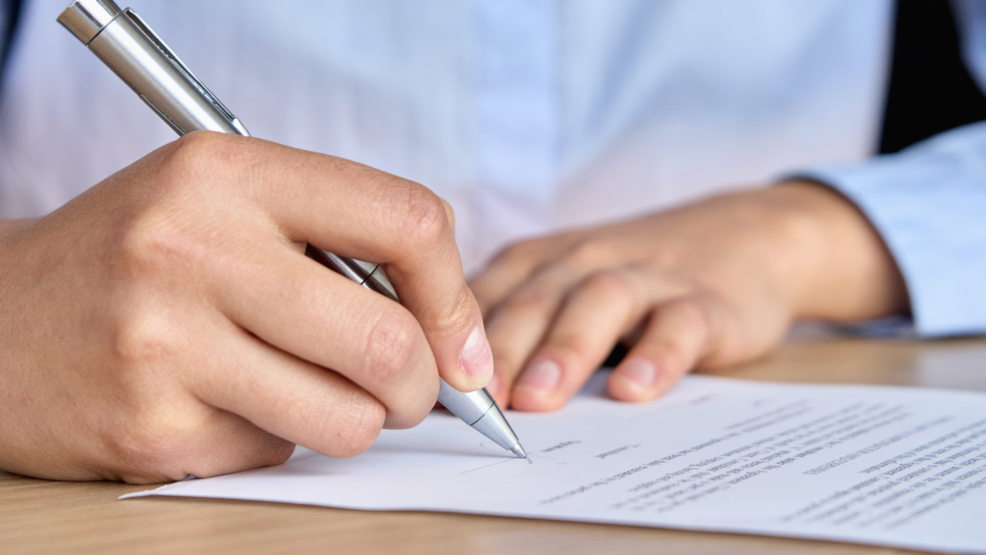 A person wearing a blue shirt is signing a piece of paper with a silver pen.