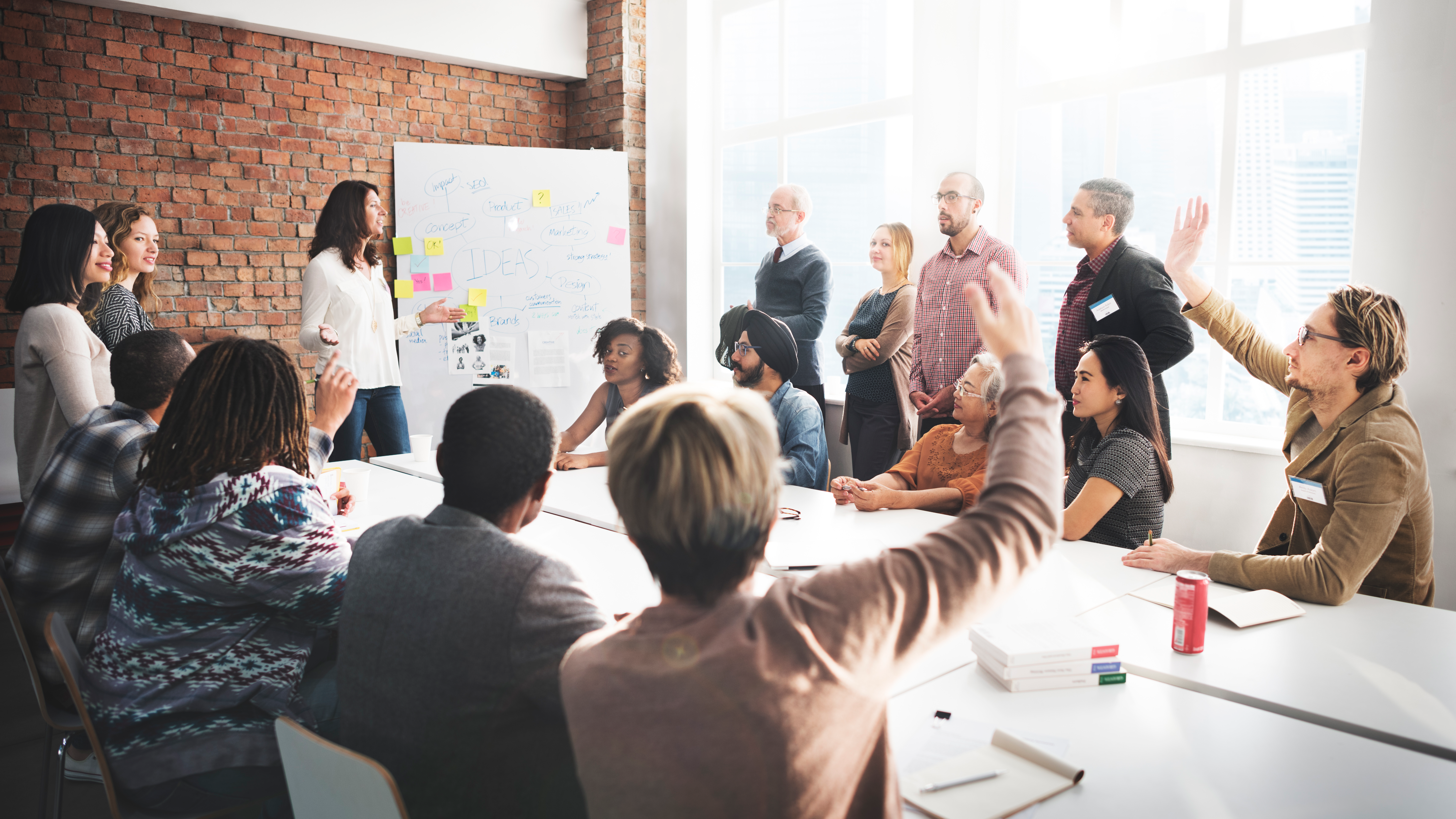 A woman standing next to a white board with notes on leads a discussion with a group of 15 adult learners sitting and standing around a table.