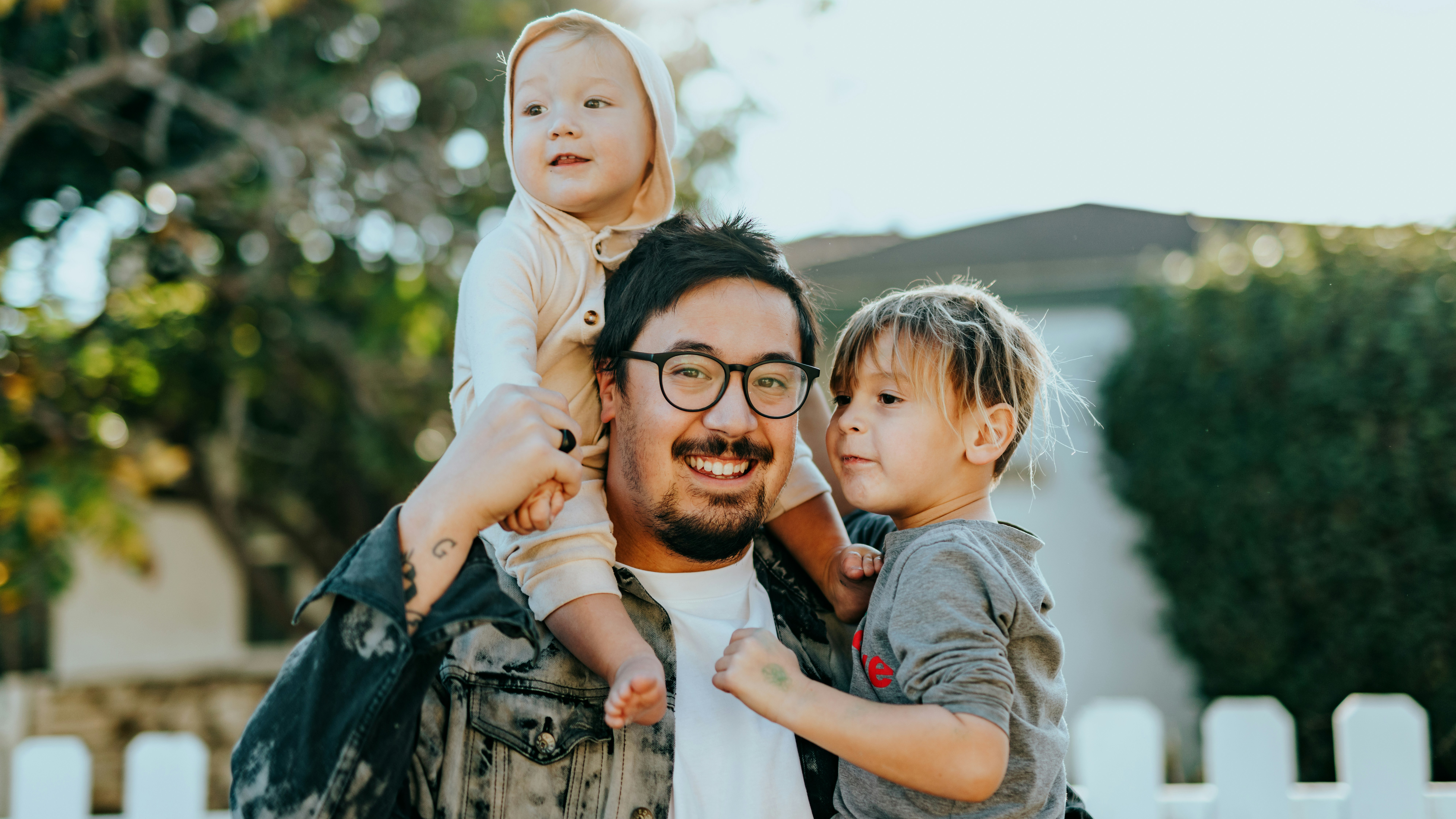 A smiling man with glasses and a goatee stands outside with one child sitting on his shoulders and holding another child in his arm.