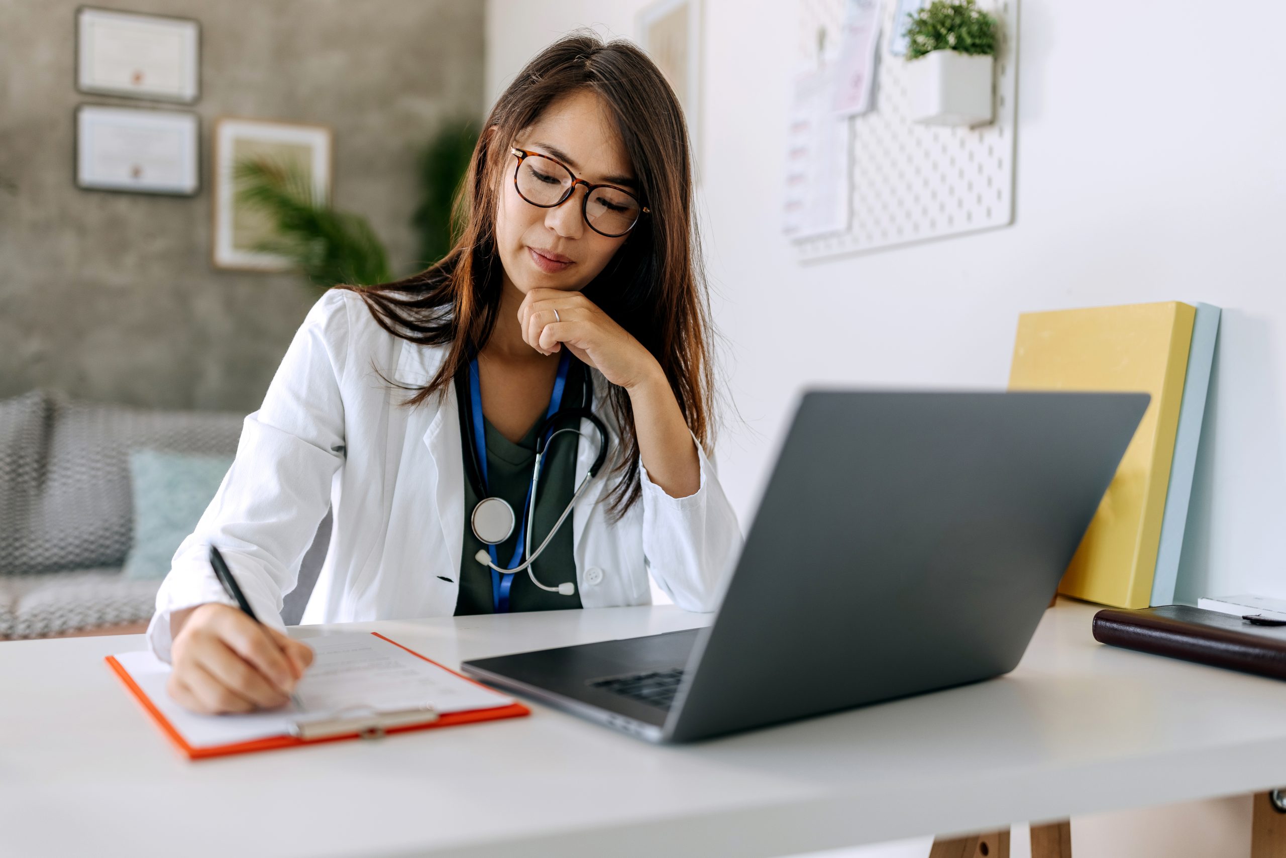 A female clinician looks at a laptop and takes notes on a clipboard.