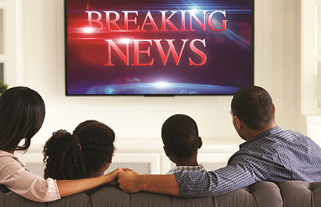 A family on their living room sofa watching the TV. The TV screen says Breaking News.