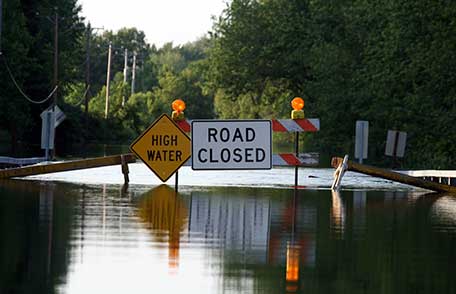 A flooded residential street with High Water and Road Closed signs blocking entry.
