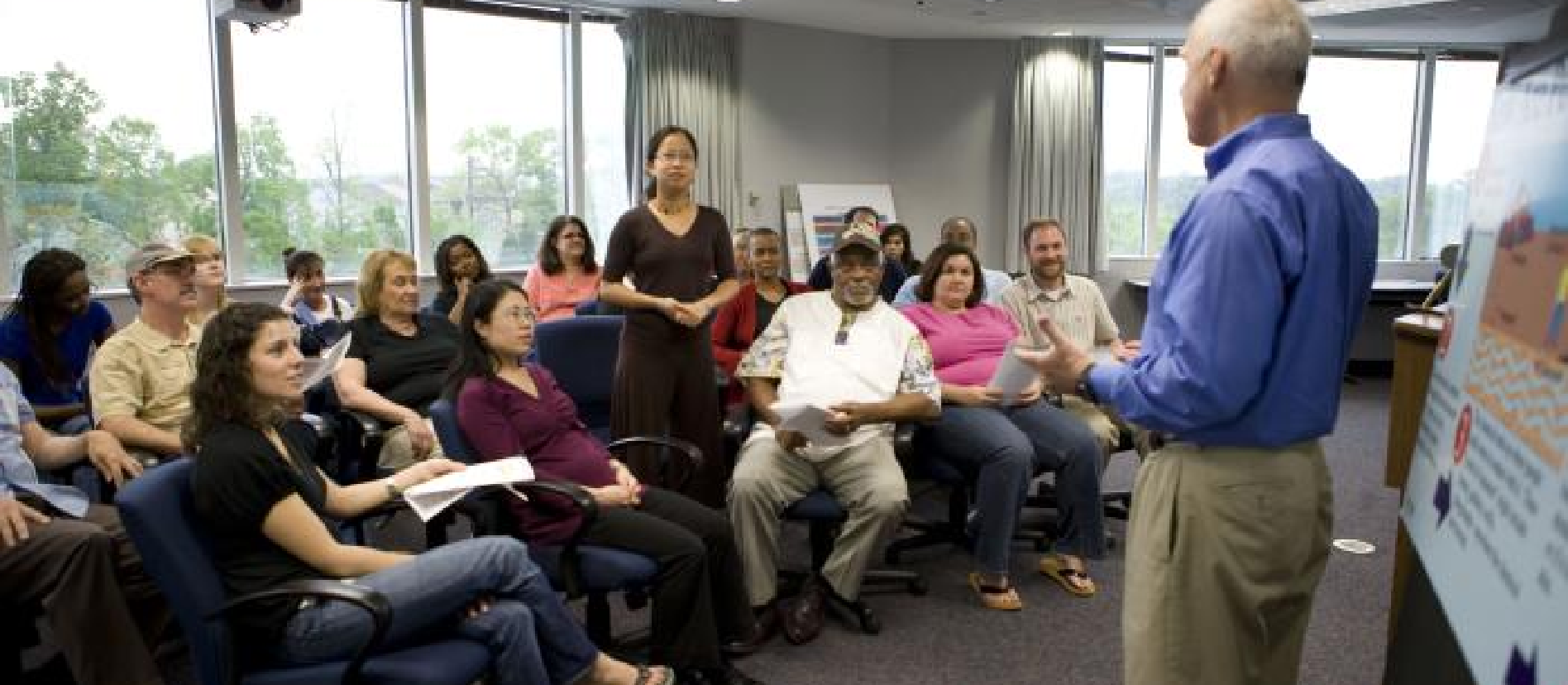 A woman standing to ask the male presenter a question with white hair and a blue shirt in front of a group of attendees.