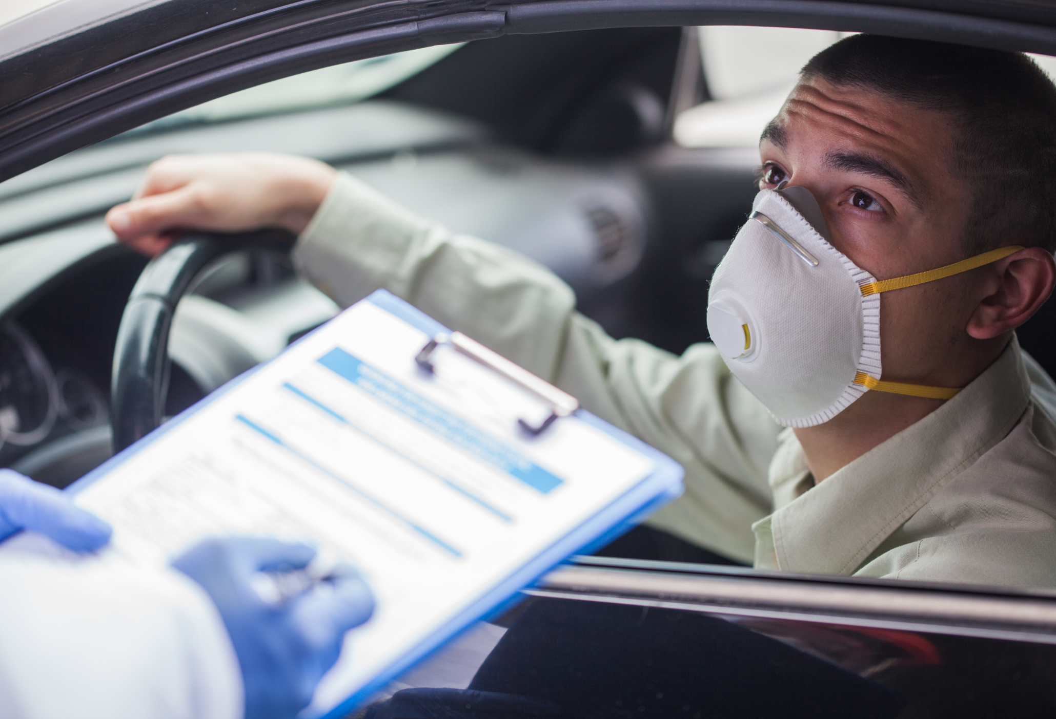 A man wearing a white mas is looking up at a person with a clipboard at a drive-up testing event for COVID-19.