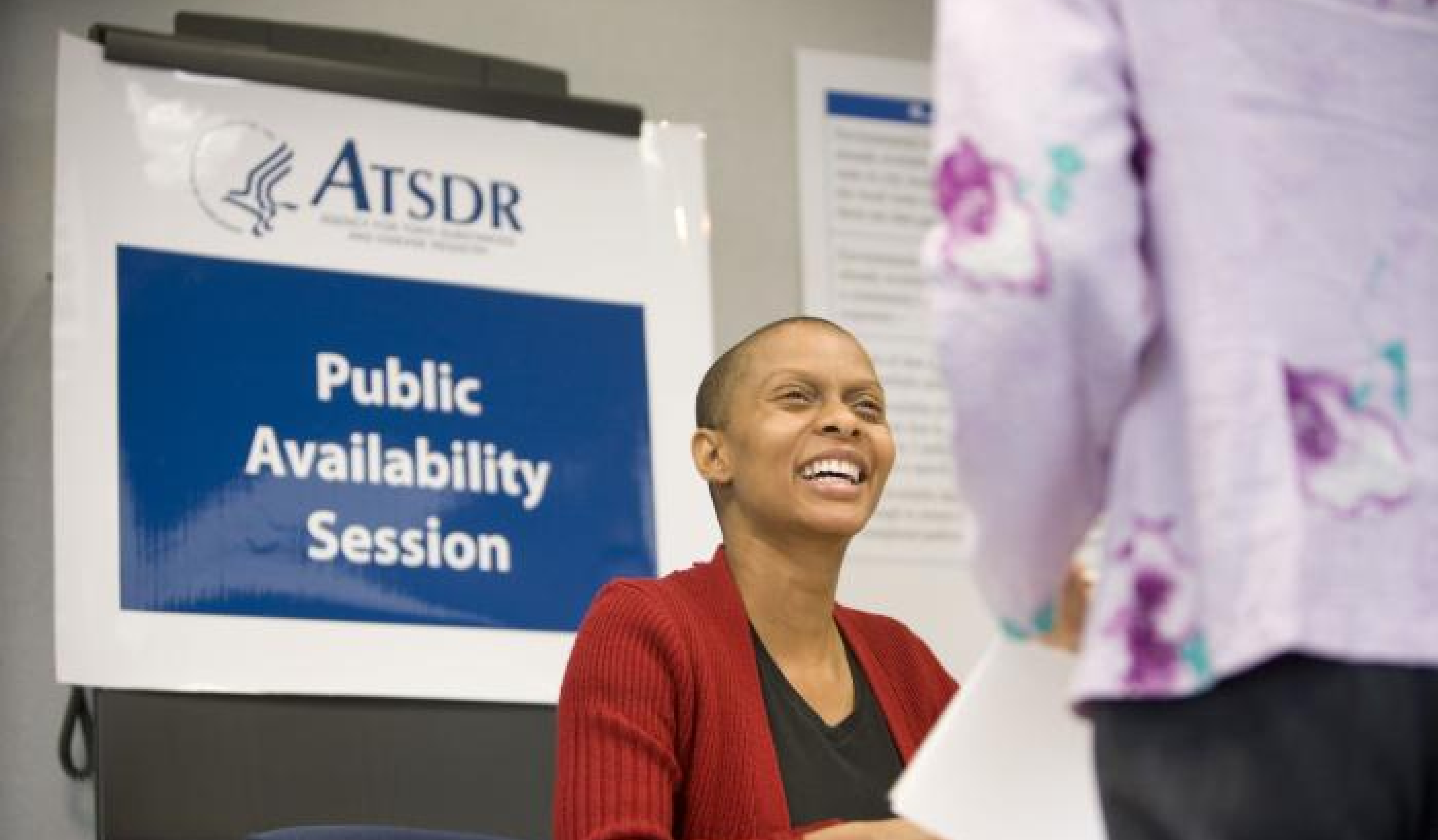 A woman in a red sweater and black shirt is smiling up at a person with a light purple sweater in from of an ATSDR session sign.