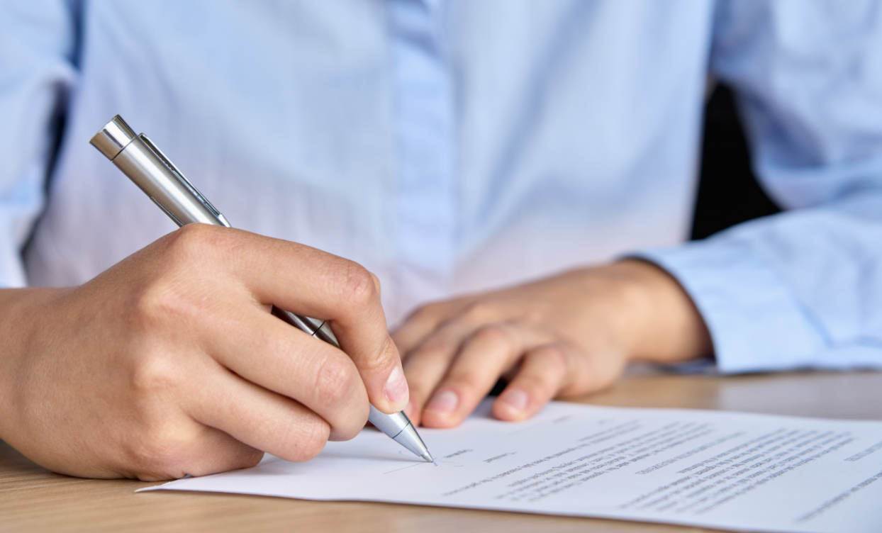 A person in a blue shirt is holding a silver pen writing on a sheet of paper.