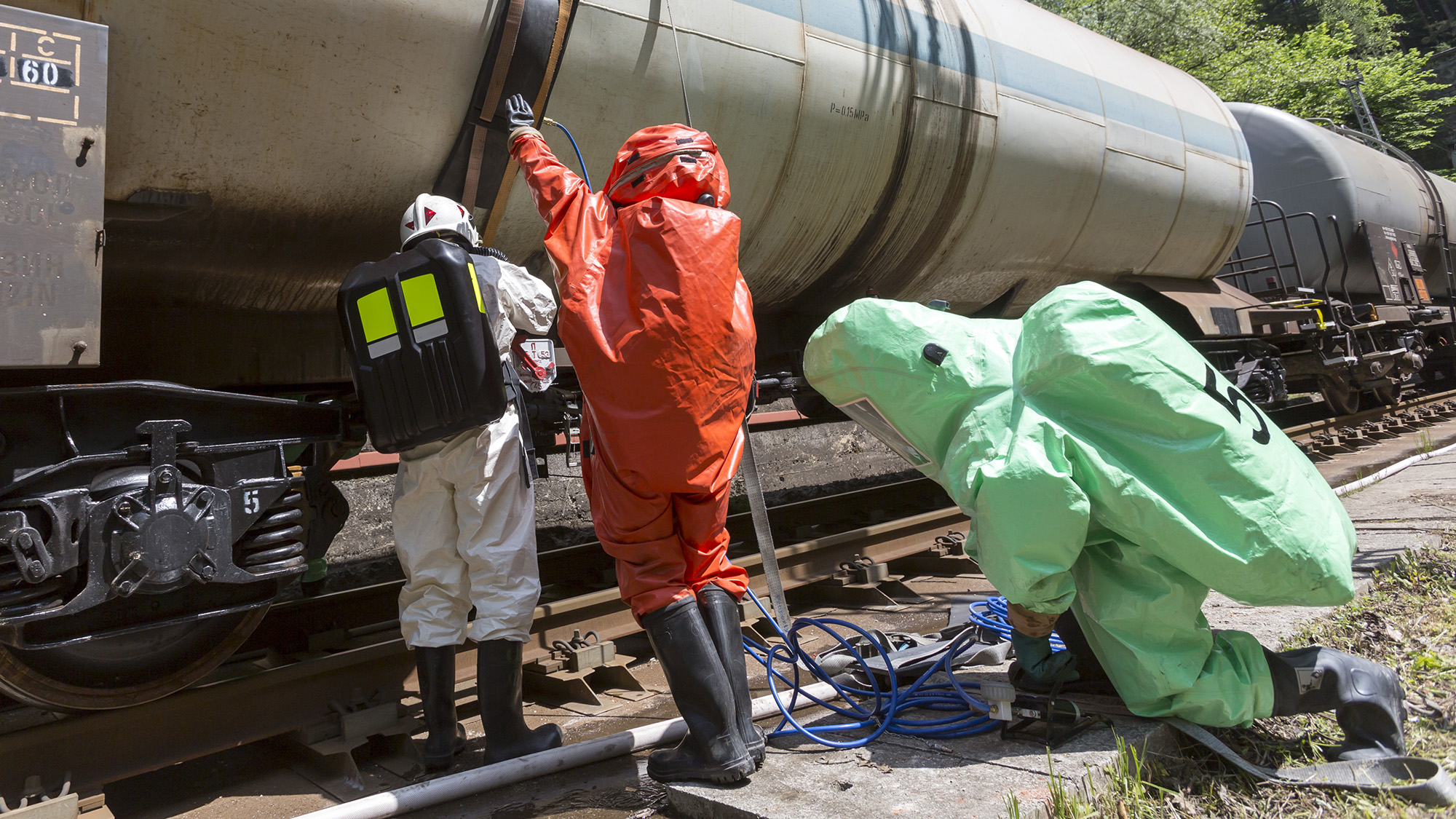 Three workers in hazmat suits inspect a train tanker car.