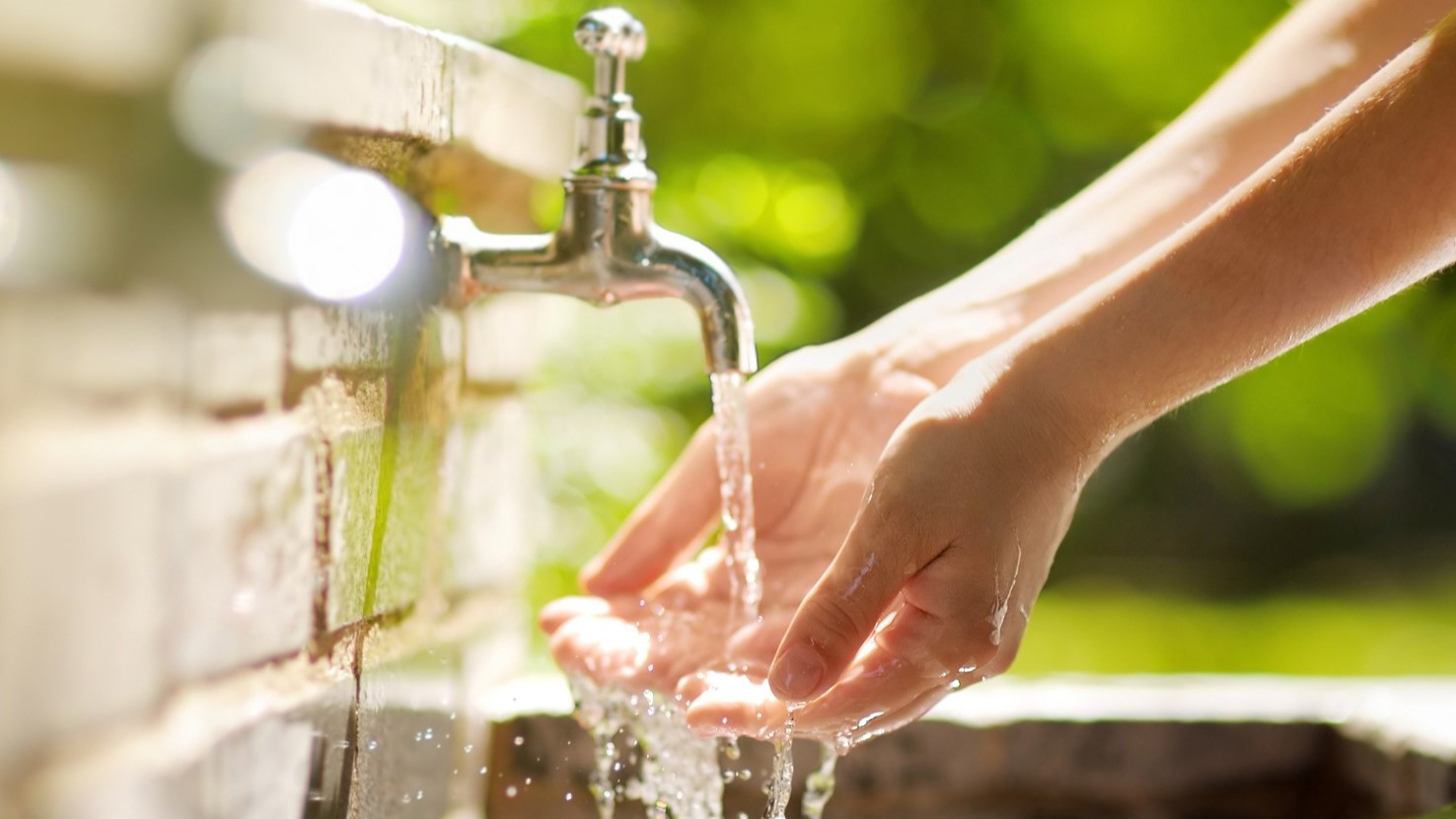 Two hands washing in an outdoor sink made of brick with greenery in the background.