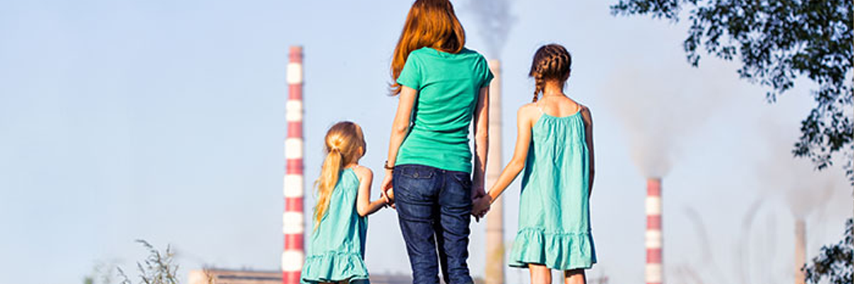 Woman holds hands with two children while looking at a factory that has two red and white smokestacks