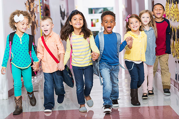 Seven young children walk in a hallway holding hands and smiling. Some are wearing backpacks.