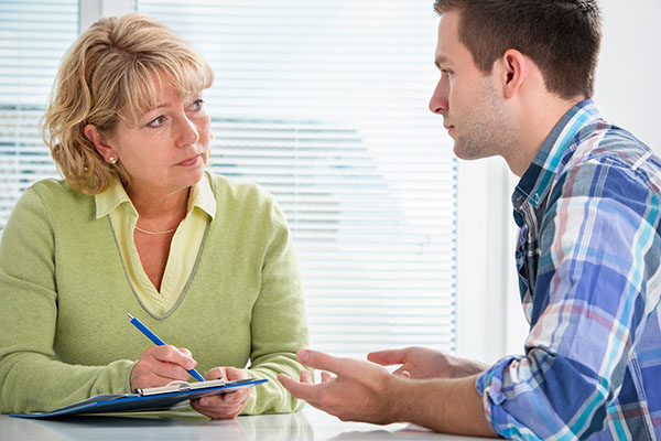 Young white man in blue speaks to a blonde white woman in green, who is taking notes