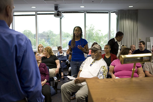 A woman in a blue shirt addresses a crowd of people at a meeting
