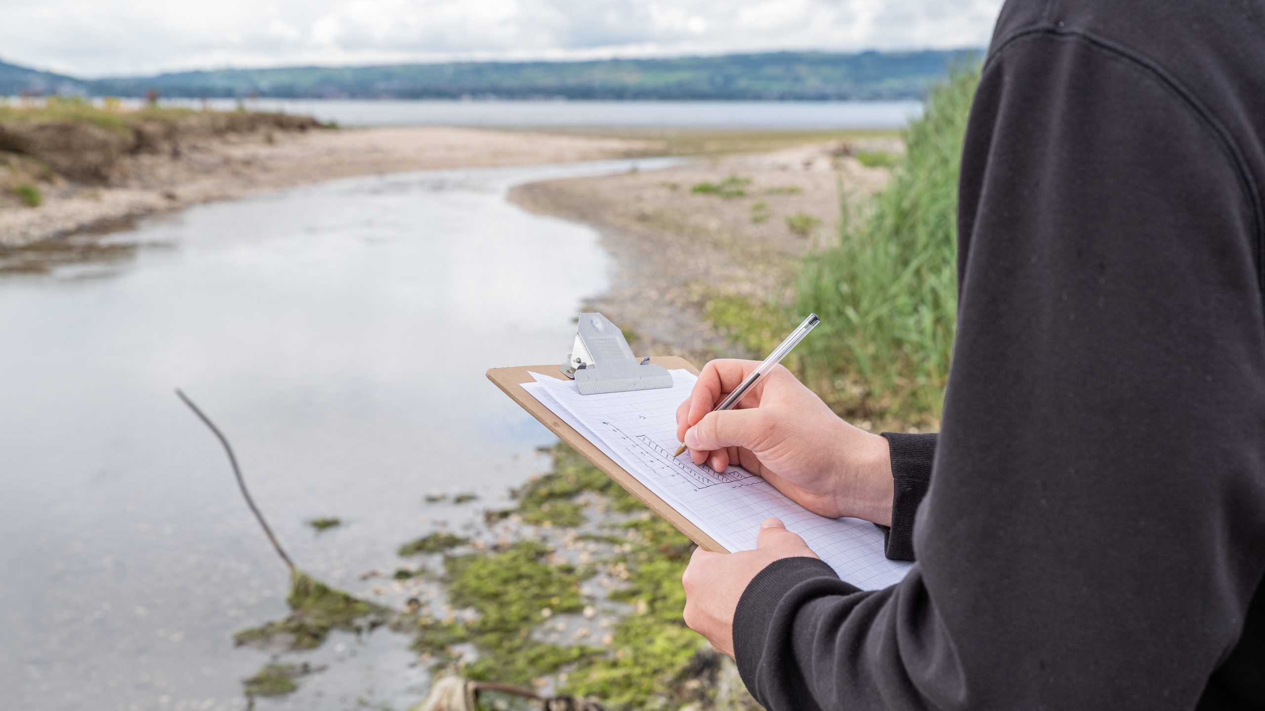 A man is making observations of a river and coastline with a clipboard.